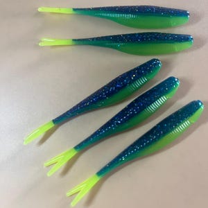 May include: Five green and blue fishing lures with glitter. The lures have a split tail and a pointed head.