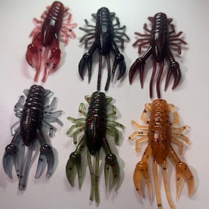 May include: Six crawfish-shaped fishing lures in various colors, including red, black, green, and orange. The lures have detailed sculpting and realistic colors.