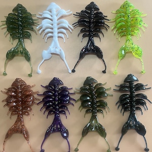 May include: Assortment of eight soft plastic fishing lures in various colors, including green, white, black, brown, purple, and blue. The lures have a segmented body with a segmented tail.