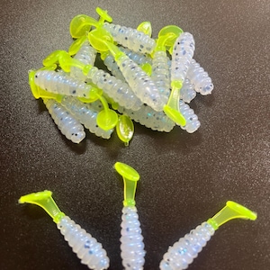 May include: A bulk pack of white and blue speckled fishing lures with neon green tails. The lures are in a pile and three are separated on a black surface.