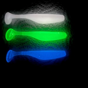 May include: Three soft plastic fishing lures with paddle tails arranged vertically. The top lure is translucent white, the middle lure is vibrant neon green, and the bottom lure is bright blue. Each lure features a rounded head and a flat tail.