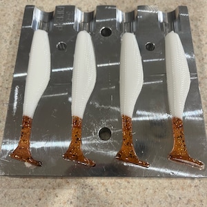 May include: Four white and brown soft plastic fishing lures in a metal mold. The lures are shaped like fish and have a textured surface.