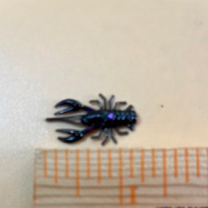 May include: A small black crawfish lure with a purple glitter accent. The lure is about 1 inch long and is shown next to a ruler.