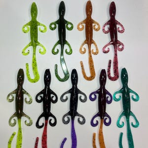 May include: Assortment of ten plastic lizard lures in various colors, including green, black, brown, purple, and teal. Each lure has a curled tail and four legs with small hooks.
