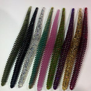 May include: A collection of nine soft plastic fishing lures, each featuring a segmented, worm-like body with a ribbed texture. The lures are presented in a variety of colors including olive, black, clear with glitter, green, pink, purple, and gold.