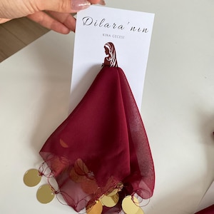 Puede incluir: Un pañuelo burdeos con adornos de monedas doradas. El pañuelo está sujeto a una tarjeta blanca con el texto "Dilara'nın" y "KINA GECESI". El pañuelo está hecho de una tela transparente.