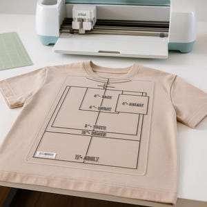 May include: A beige t-shirt with a clear plastic alignment guide for heat transfer vinyl. The guide has black lines and labels for different shirt sizes, including "Adult" and "Youth". A Cricut machine is in the background.