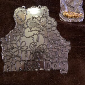 May include: A silver metal cutout of a cartoon bear holding flowers with the text "Have a Nice Day" around it. The cutout has two holes at the top for hanging.