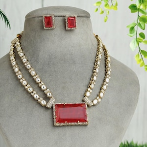 May include: A gold-toned necklace and earring set. The necklace features a large, rectangular red gemstone pendant, accented with smaller white stones and a double-strand design. Matching square earrings with red gemstones complete the set.