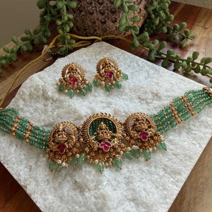 May include: A gold-toned necklace and matching earrings set. The necklace features three ornate pendants with red and green accents, connected by strands of green beads. The earrings mirror the pendant design. The jewelry is displayed on a white surface.
