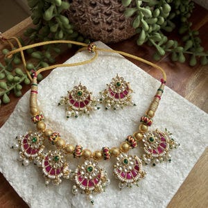 May include: A gold-toned necklace and earring set. The necklace features a series of ornate, circular pendants with red and green accents, and small white pearls. The earrings mirror the necklace design. The jewelry is displayed on a white surface.