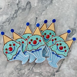 May include: A group of six blue, green, and yellow embroidered iron-on patches featuring a smiling ice cream cone with the text "The Soft Scoop" on each.