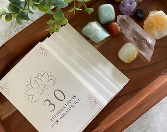 May include: A stack of white cards with the text "30 Affirmations for Abundance" on the top card. The cards are in a wooden bowl with various crystals and stones, including amethyst, clear quartz, and citrine.