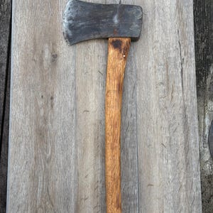 May include: A vintage axe with a weathered steel head and a wooden handle. The axe head shows signs of wear and use. The wooden handle is a light brown color. The axe is resting on a light gray wooden surface.