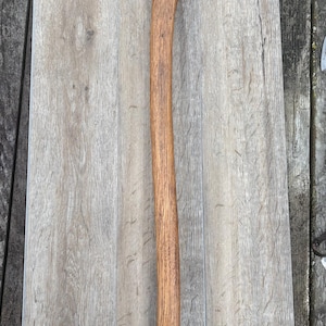 May include: An axe with a dark metal head and a long, curved wooden handle. The handle is a light brown color, and the axe head is a darker gray. The axe is lying on a light gray wooden surface.