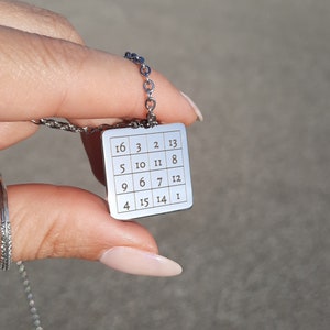 Könnte beinhalten: Eine silberne Halskette mit einem quadratischen Anhänger, der ein nummeriertes Gitter zeigt. Die Zahlen auf dem Anhänger sind 16, 3, 2, 13, 5, 10, 11, 8, 9, 6, 7, 12, 4, 15, 14 und 1.