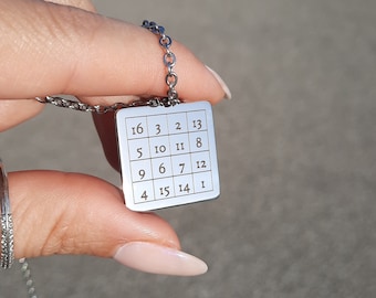 May include: A silver necklace with a square pendant featuring a numbered grid. The numbers on the pendant are 16, 3, 2, 13, 5, 10, 11, 8, 9, 6, 7, 12, 4, 15, 14, and 1.
