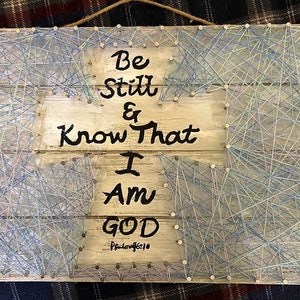 May include: A wooden cross with blue string art and the words "Be Still & Know That I Am God - Psalm 46:10" written on it.