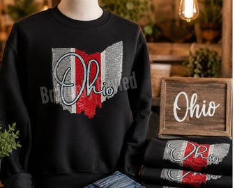 May include: A black long-sleeve sweatshirt featuring a sequined graphic of the state of Ohio in silver and red, with the word Ohio written in white script across the center. The design is repeated on a stack of folded black sweatshirts nearby.
