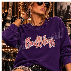 May include: A deep purple long-sleeve crewneck sweatshirt featuring the word Bulldogs in a red, glittery, cursive font with a white outline. The garment has a relaxed fit and a solid, dark purple fabric construction throughout the body.