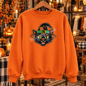 May include: An orange long-sleeve sweatshirt featuring a graphic print of a black cat wearing a witch hat. The hat is decorated with a green band, a buckle, a crescent moon, and a flower. The cat has bright green eyes and visible fangs.