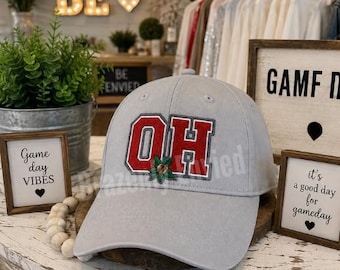 May include: A light gray baseball cap featuring a red and white embroidered patch with the letters OH and a small green leaf accent. The cap has a structured crown, a curved brim, and stitched eyelets on the top panels for ventilation.