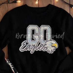 May include: Black crew neck sweatshirt with the words "GO Eagles" in silver sequin lettering. An eagle head graphic is on the right side of the text. The sweatshirt is on a wooden surface.