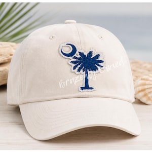 May include: A cream-colored baseball cap featuring an embroidered navy blue palmetto tree and crescent moon design on the front panel. The cap has a structured crown, a curved brim, and a button on top, with the text BrazenlyEnvied overlaid.