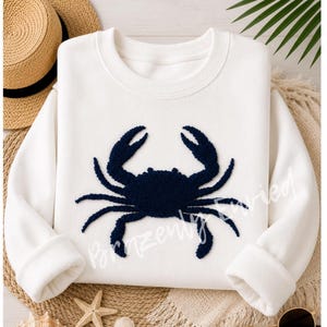 May include: A white long-sleeve sweatshirt featuring a dark navy blue textured crab applique centered on the front. The sweatshirt has a crew neckline and ribbed cuffs. The text Brazenly Envied is written in a light script across the crab graphic.