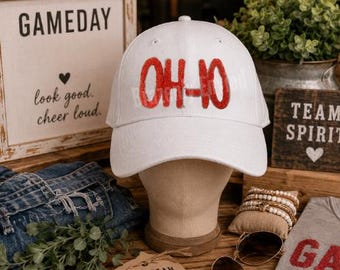 May include: A white baseball cap featuring the text OH-IO in red, textured, sequin-style lettering across the front panel. The cap has a structured crown, a curved brim, and eyelets for ventilation, presented on a neutral-toned display head.
