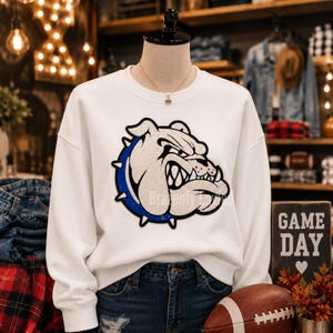 May include: A white long-sleeve sweatshirt featuring a large, central graphic of a bulldog head with a blue spiked collar. The bulldog design is rendered in white and black with a textured appearance. The sweatshirt has a crew neckline and long sleeves.