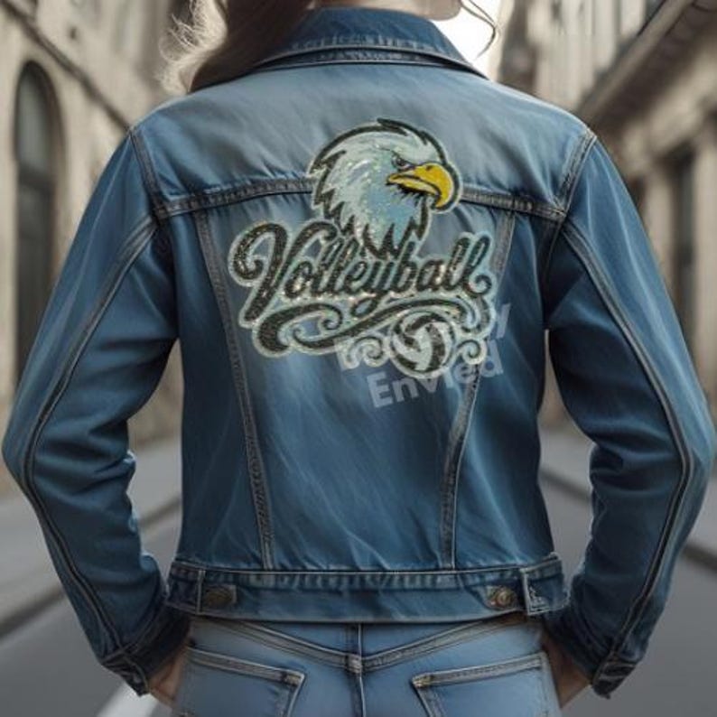 Pu&ograve; includere: Una giacca di jeans blu con una grande grafica sul retro che raffigura una testa di aquila calva sopra la parola Volleyball in carattere corsivo. Il design include una piccola icona di pallavolo e la parola Envied in un carattere sans-serif.