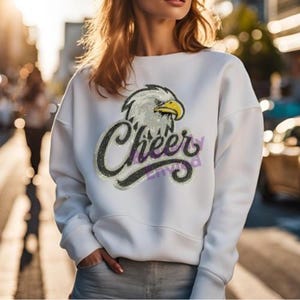 May include: A white long-sleeve sweatshirt featuring a graphic of a bald eagle head with a yellow beak positioned above the word Cheer in a black cursive script. The design includes a subtle glittery texture throughout the eagle and text.