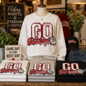 May include: A white crewneck sweatshirt featuring the text GO Bulldogs in red and silver sequins with a bulldog graphic. The design is repeated on folded white, gray, and black sweatshirts, and a black baseball cap with Bulldogs text.
