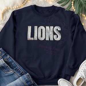 May include: Navy blue sweatshirt with the word "LIONS" in a textured, silver-gray font. The text "Brazenly Envied" is below the word "LIONS". The sweatshirt is paired with blue jeans and white sneakers.
