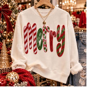 May include: A white long-sleeve crewneck sweatshirt featuring the word Merry in large, colorful, rhinestone-embellished letters. The letters are decorated with red and white candy cane stripes, green and red polka dots, and green stripes.