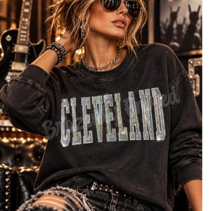 May include: A charcoal grey long-sleeve crewneck sweatshirt featuring the word CLEVELAND printed in large, block-style letters with a reflective, multi-colored holographic glitter finish across the chest area of the garment.