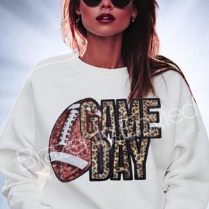 May include: White sweatshirt with a football and "GAME DAY" text. The football is brown and red with white stitching. The text has a leopard print design. A stylish and comfortable top for game day.