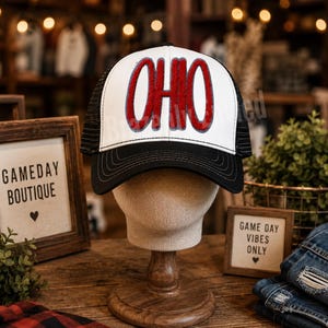 May include: A trucker hat with a white front panel featuring the word OHIO in red chenille letters with a grey outline. The hat has a black brim with decorative stitching and black mesh side and back panels, displayed on a mannequin head.