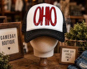 May include: A trucker hat with a white front panel featuring the word OHIO in red chenille letters with a grey outline. The hat has a black brim with decorative stitching and black mesh side and back panels, displayed on a mannequin head.
