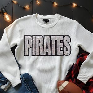 May include: White sweater with the word "PIRATES" in silver sequins. The sweater is paired with blue jeans, a red and black plaid shirt, and a football. Decorative string lights are in the background.