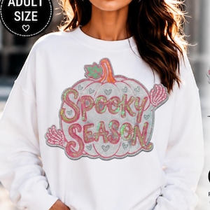 May include: A white long-sleeve sweatshirt featuring a large graphic of a pumpkin with the text "SPOOKY SEASON" in iridescent pink and green lettering. The design includes small heart shapes and skeleton hand accents on the sides.