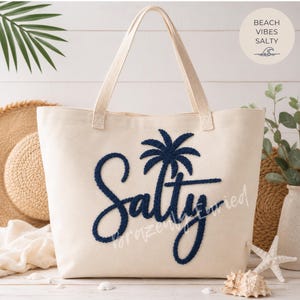 May include: A cream-colored canvas tote bag featuring the word Salty in dark navy blue textured embroidery, with a palm tree icon replacing the letter t. The bag has two sturdy fabric handles and a clean, minimalist design for daily use.