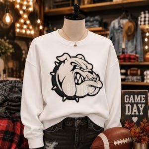 May include: A white long-sleeve sweatshirt featuring a large, central graphic of a snarling bulldog head in black and white. The bulldog design is detailed with sharp teeth and a spiked collar. The sweatshirt has a crew neckline and long sleeves.