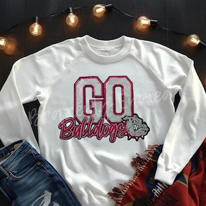 May include: White sweatshirt with the text "GO Bulldogs" in a pink and silver design. A bulldog graphic is below the text. The sweatshirt is paired with blue jeans and a red and blue patterned blanket.