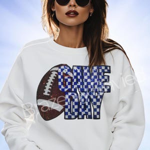 May include: White sweatshirt featuring a football graphic and the words "GAME DAY" in a blue and white checkered pattern. The football is brown with white stripes. A casual, sporty design.