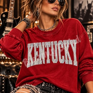 May include: A red long-sleeve crewneck sweatshirt featuring the word KENTUCKY printed in large, silver, glittery block letters across the chest. The garment has a slightly faded, vintage-style wash and a ribbed collar, cuffs, and hem.