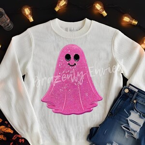May include: White long-sleeved sweater featuring a large, glittery pink ghost graphic with a smiling face. The ghost design is centered on the front of the sweater. The sweater is paired with blue denim jeans.