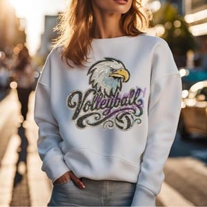 Pu&ograve; includere: Una felpa bianca a maniche lunghe con una grafica centrale raffigurante una testa di aquila calva sopra la parola Volleyball in corsivo, accentuata da una piccola icona di pallavolo. Il design &egrave; realizzato con paillettes scintillanti.