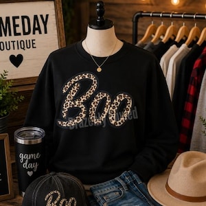 May include: A black long-sleeve sweatshirt featuring the word Boo in a brown and black leopard print pattern with a dark outline. The sweatshirt has a crew neckline and is displayed on a mannequin wearing a gold chain necklace.